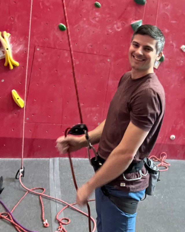 A team member smiling at an indoor climbing wall, holding a belay rope